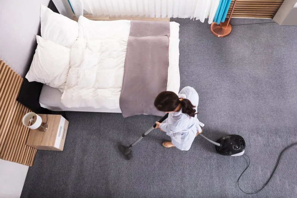 An overhead shot of a person in a blue robe vacuuming a grey carpet next to a neatly made bed. The room appears to be a bedroom with a bedside table holding a lamp and a decorative wooden wall panel. Curtains and a floor lamp are visible in the background, suggesting a domestic setting.