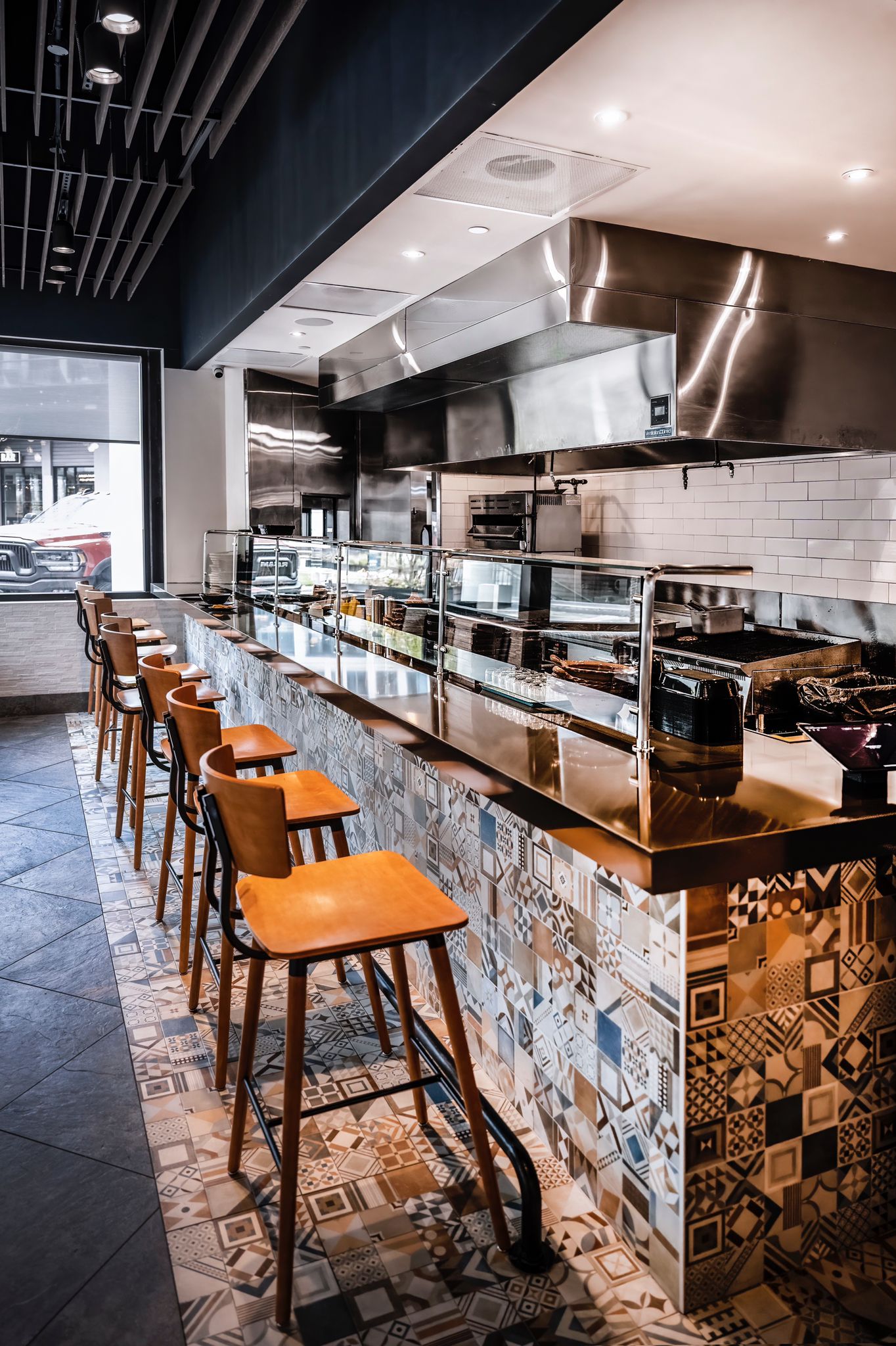 The open kitchen offers a dynamic experience, with chefs preparing meals in a stainless-steel workspace. The tiled front of the counter and warm-toned stools complement the contemporary decor.