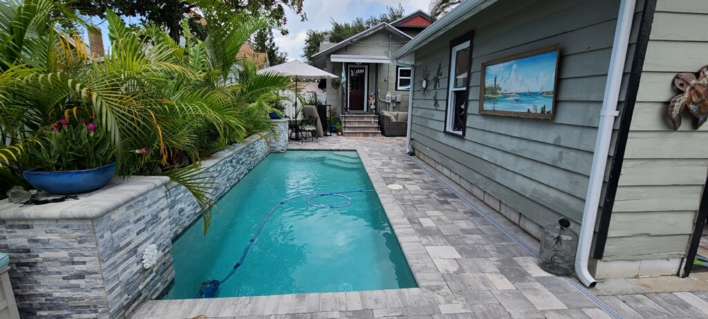 Narrow residential lap-style pool beside the home, surrounded by gray paver decking and tropical landscaping.