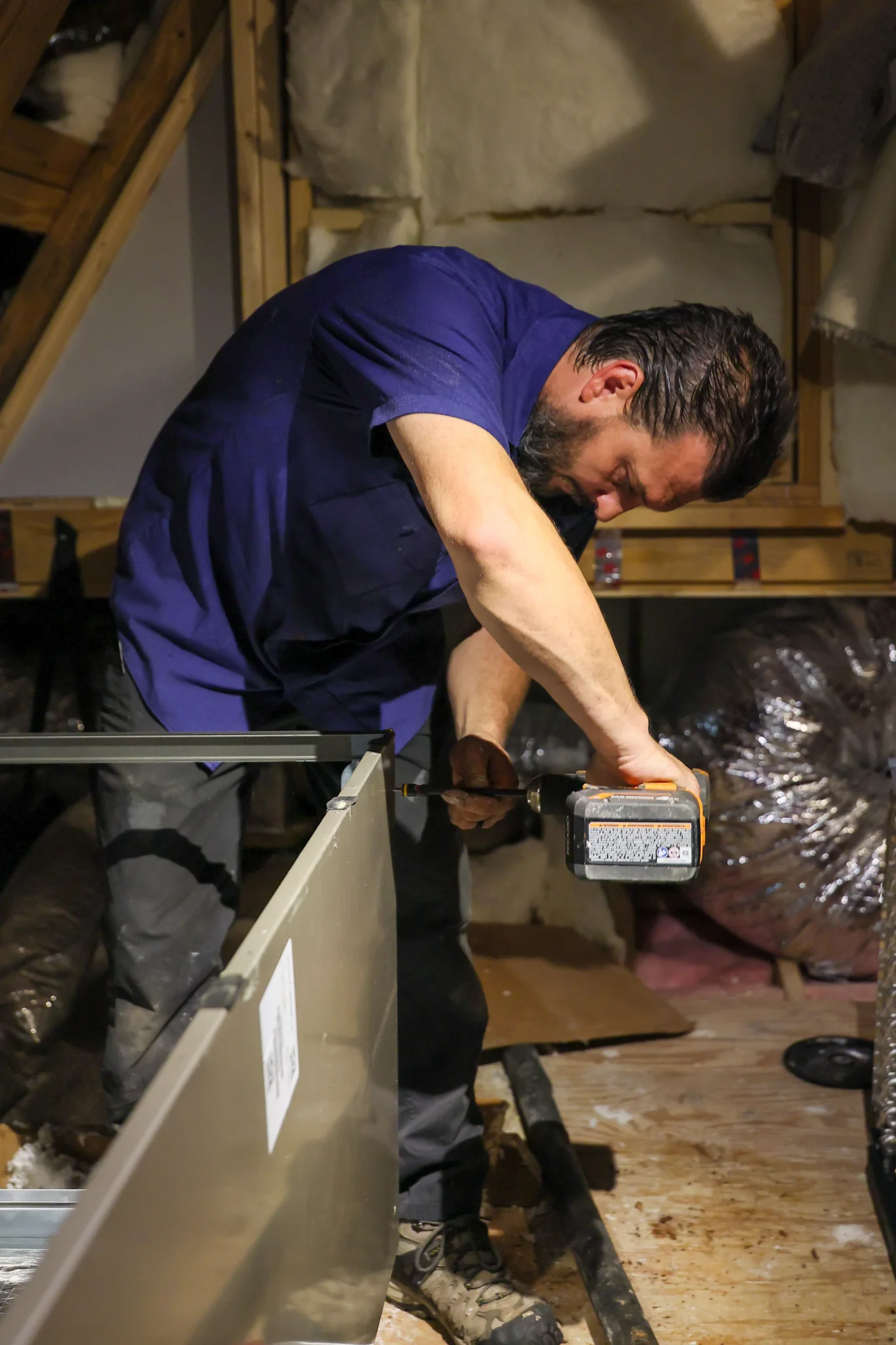 A man wearing a blue shirt and dark pants is working in an attic, using a cordless drill to screw a  