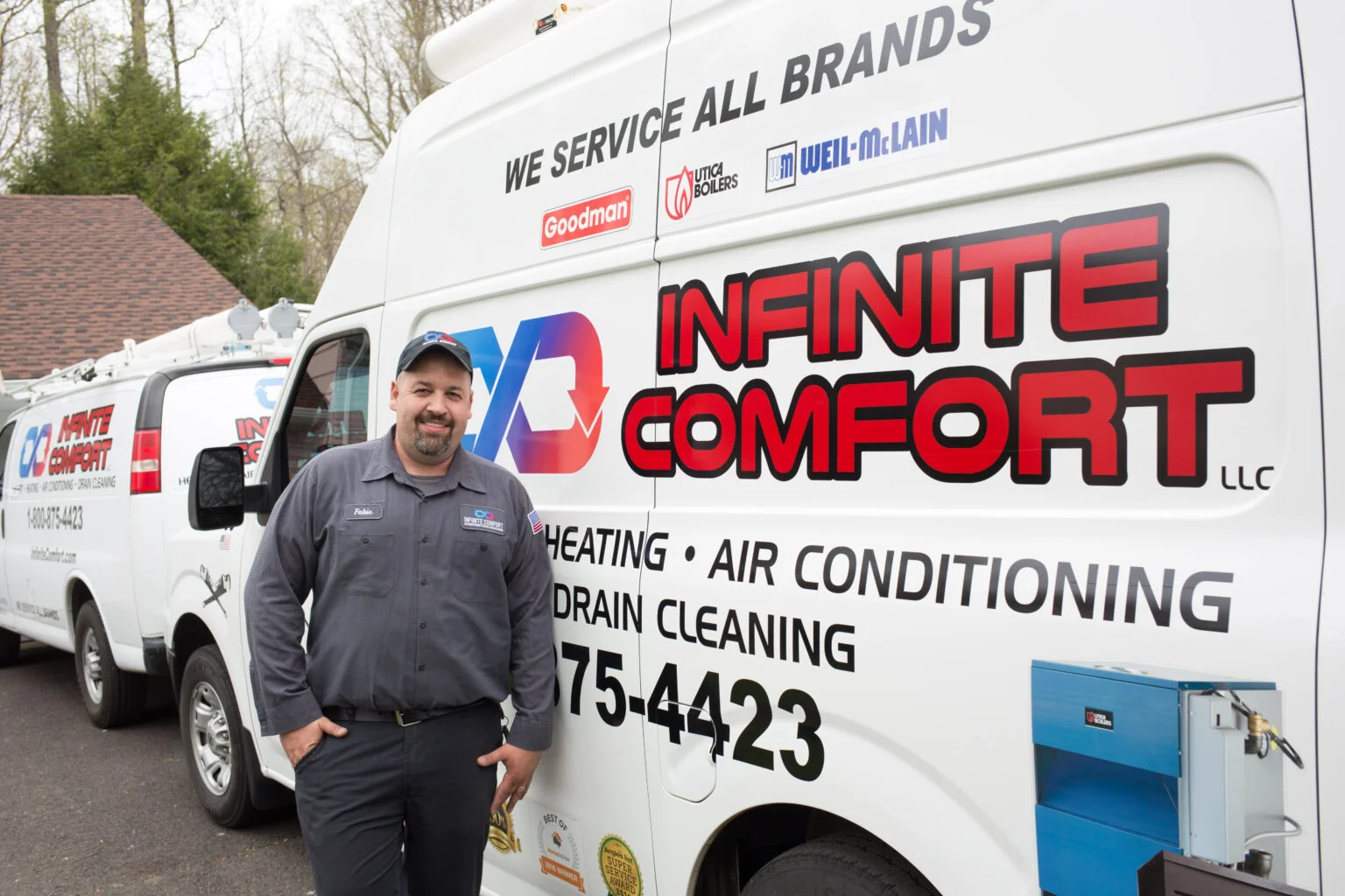 A man in a grey uniform and a baseball cap stands in front of a white commercial van, which displays the company name "INFINITE COMFORT LLC" prominently in large red lettering. The van is also labeled with services like "HEATING • AIR CONDITIONING • DRAIN CLEANING" and a phone number "75-4423". There are other vans and a house in the background. The man is smiling and has his hands in his pockets.