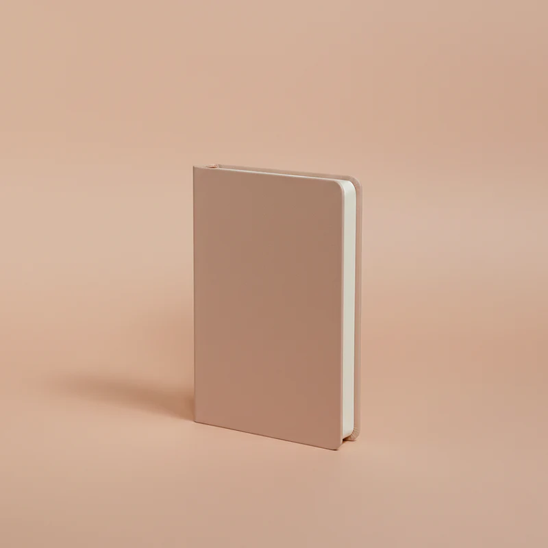 A closed, light pink, hardcover notebook stands upright on a plain, light peach-colored surface.
