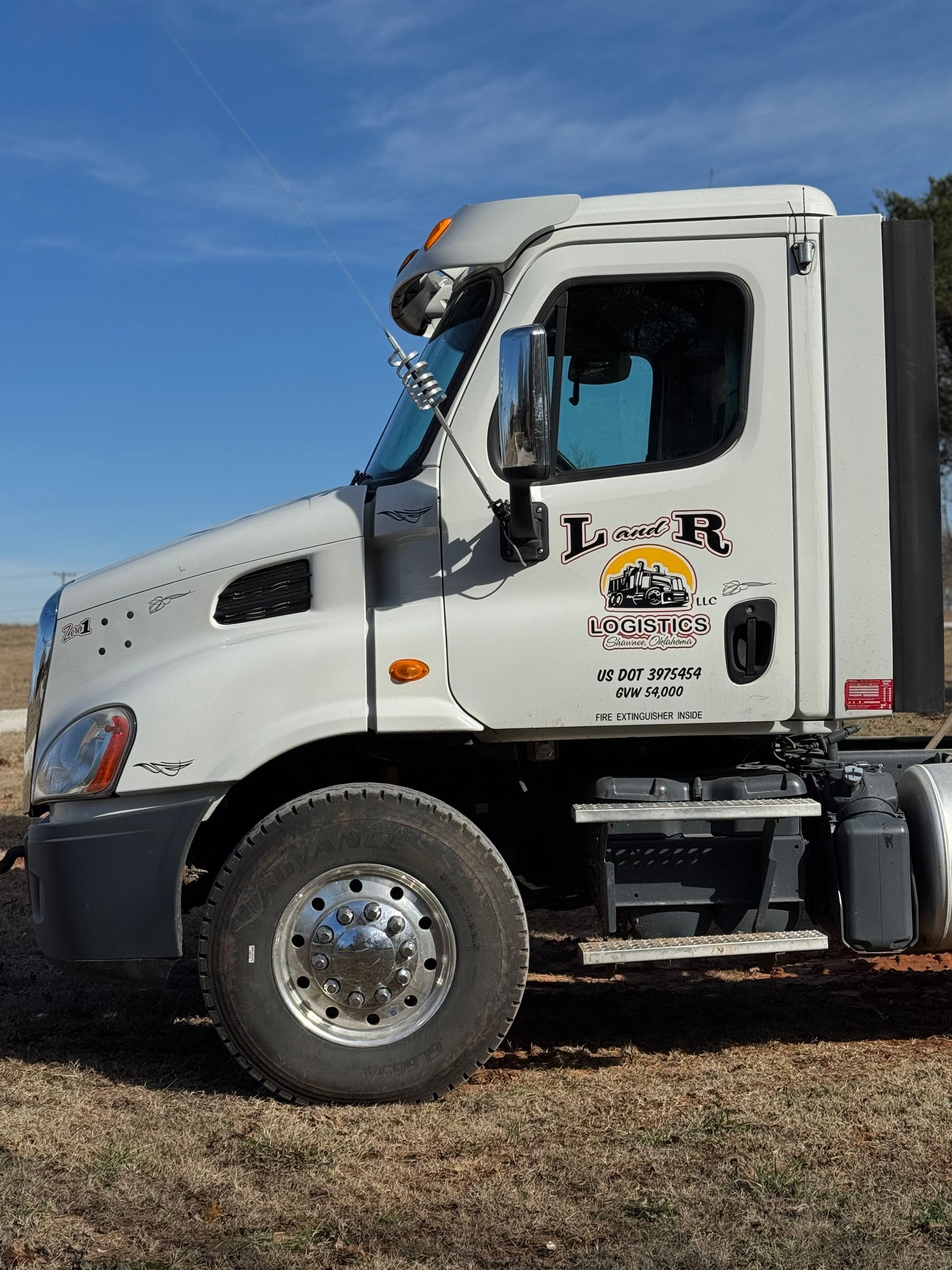 L & R Logistics LLC white semi truck photographed from the side, featuring branded cab, chrome mirror, large front tire, fuel tank, and commercial transportation equipment outdoors locally in Oklahoma.