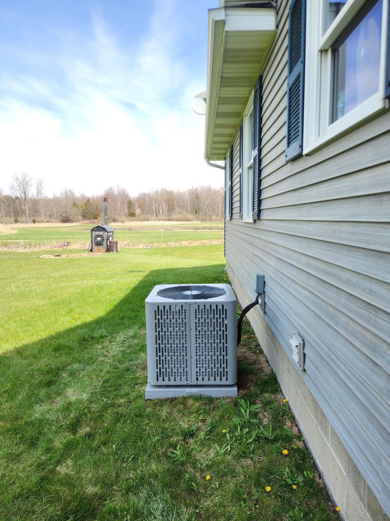 A modern grey air conditioning condenser unit is installed on a plastic pad outside a residential home. The unit sits on green grass against light grey siding under a clear sky.