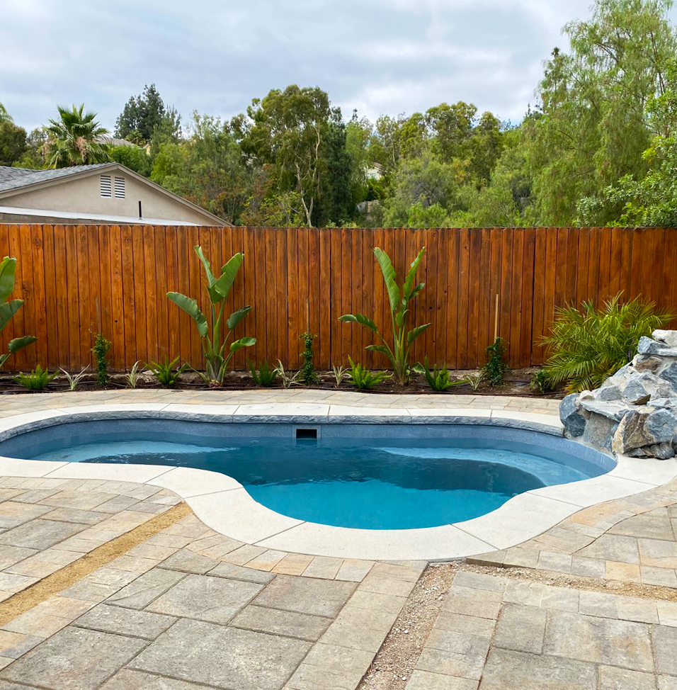 Serene backyard pool with curved blue water, stone patio, tropical plants, wooden fence, rock feature, and leafy trees under a cloudy sky.