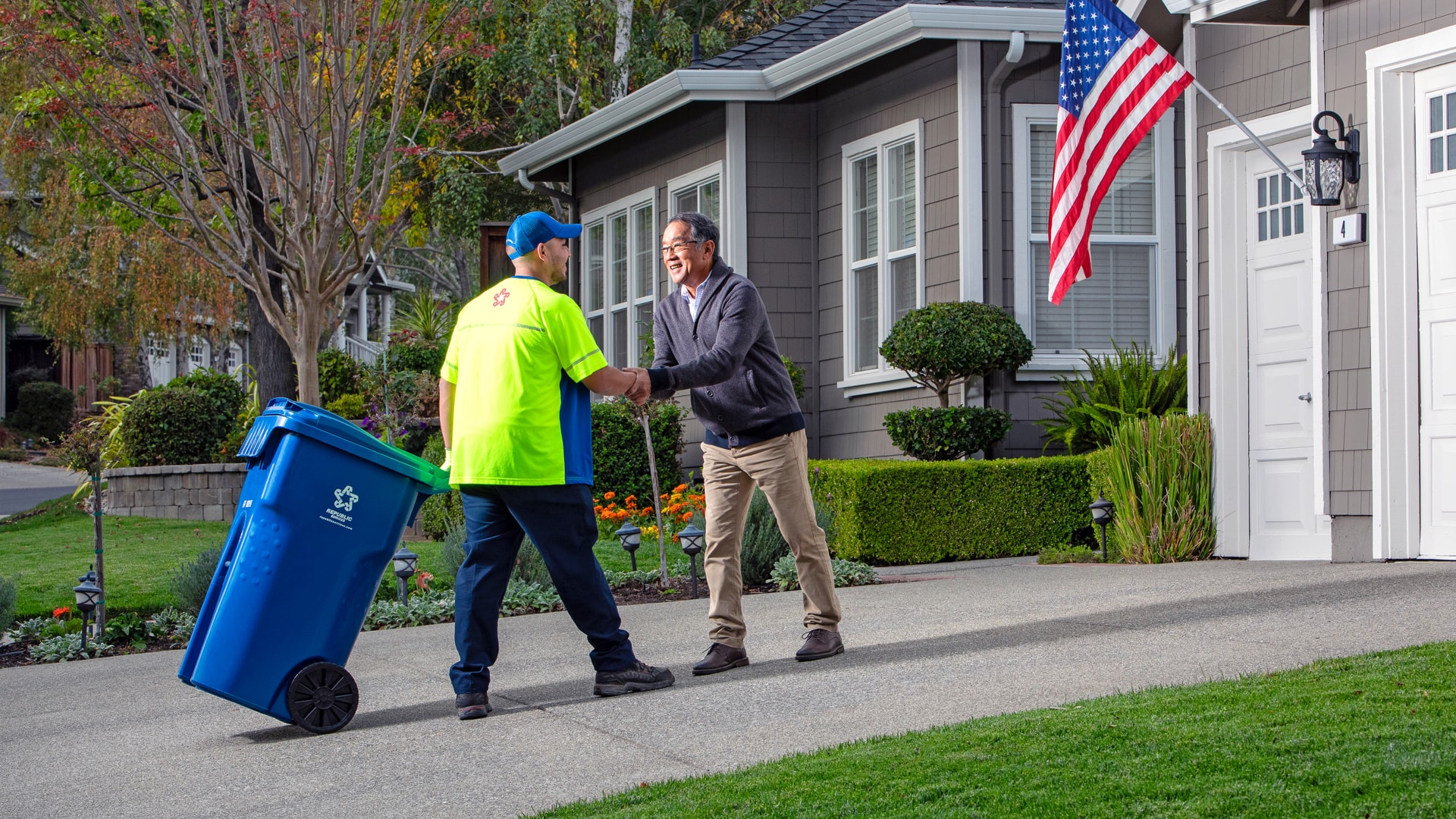 A Republic Services Employee in a bright yellow shirt rolls a blue recycling pail with the Republic Services Logo up a homeowners driveway.
