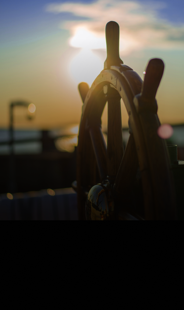 Close-up of a ship’s wooden steering wheel, with the sun setting over water in the background.