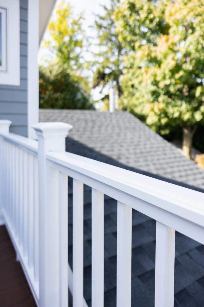 A white railing with vertical balusters runs along the edge of a deck or balcony, with a gray-sided house and a dark gray shingled roof visible behind it. Lush green trees with some yellowing autumn leaves are in the background.