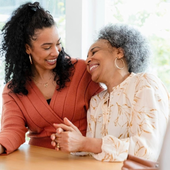 A bright, airy scene by a floor-to-ceiling window with green foliage visible outside. A younger woman in a rust-colored wrap sweater sits at a light wood table beside an older woman with silver curls in a cream floral blouse. Their hands are clasped together on the table, and they're laughing as they look at each other; the younger woman holds a phone in her free hand.