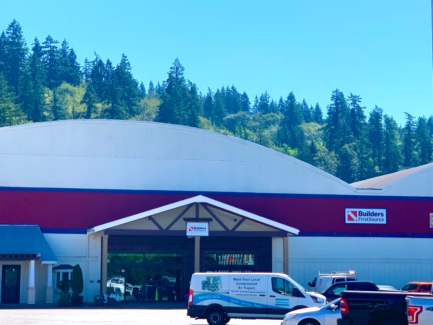 A closer look at the Builders FirstSource main entrance in Issaquah, WA, showcasing the red and white building facade, framed by evergreen trees and clear skies. Delivery vans and trucks are parked in front.