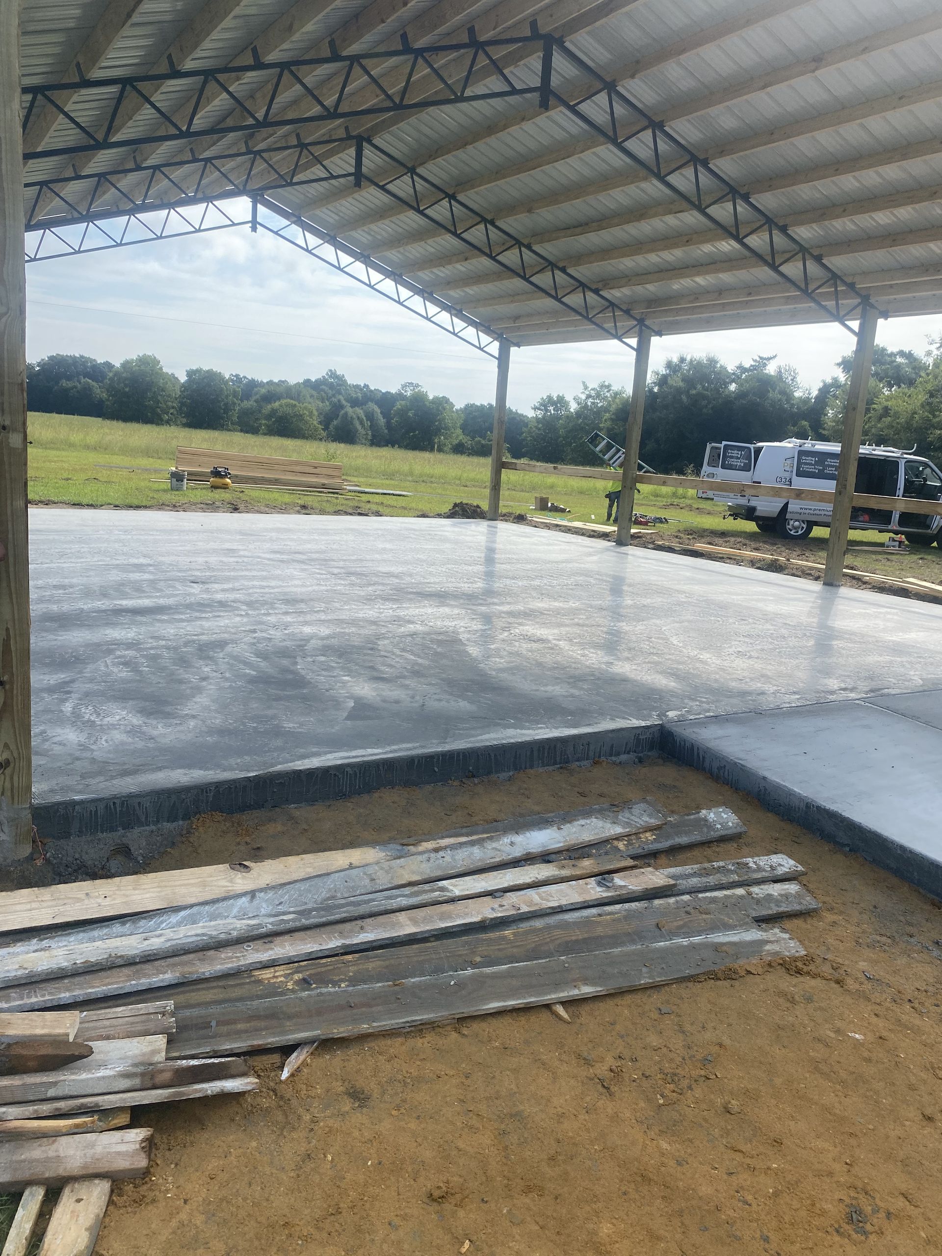 Newly poured concrete slab beneath large open-sided metal structure with exposed steel trusses and wooden posts, construction materials nearby, grassy field beyond, and work truck parked at edge.