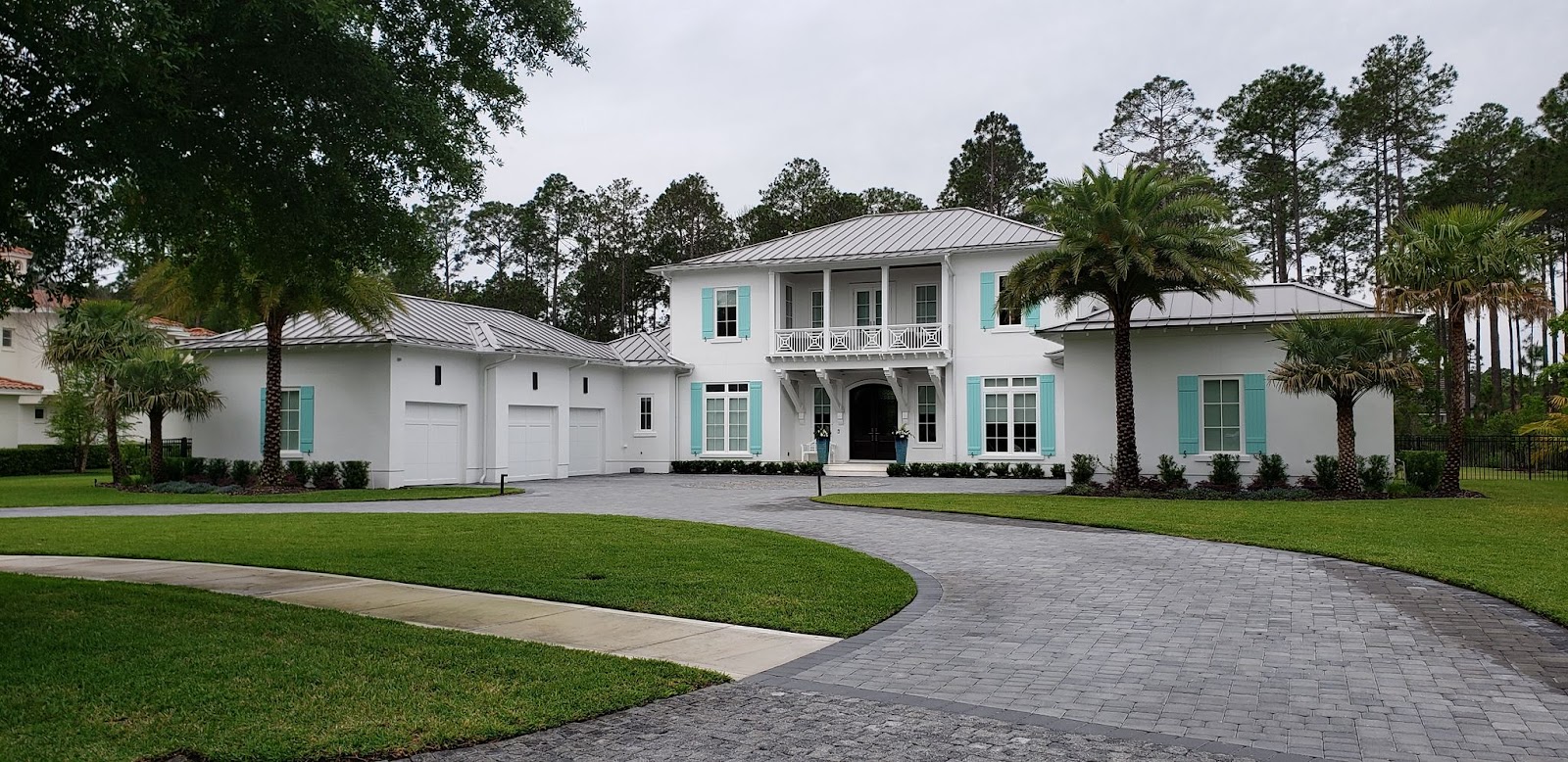 A large, modern, white house with teal shutters and a metal roof sits on a paved driveway surrounded 