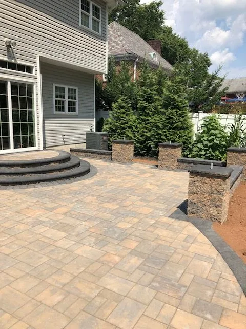 A newly constructed outdoor patio area with a large paved patio, a stone retaining wall with pillars, and a modern house in the background. The patio is made of tan and gray interlocking pavers. Curved steps with a dark border lead up to the patio from a lower level. A gray exterior wall of the house is visible with white-framed windows and a large glass door. Tall evergreen trees and other landscaping are present, along with an outdoor air conditioning unit near the base of the house.