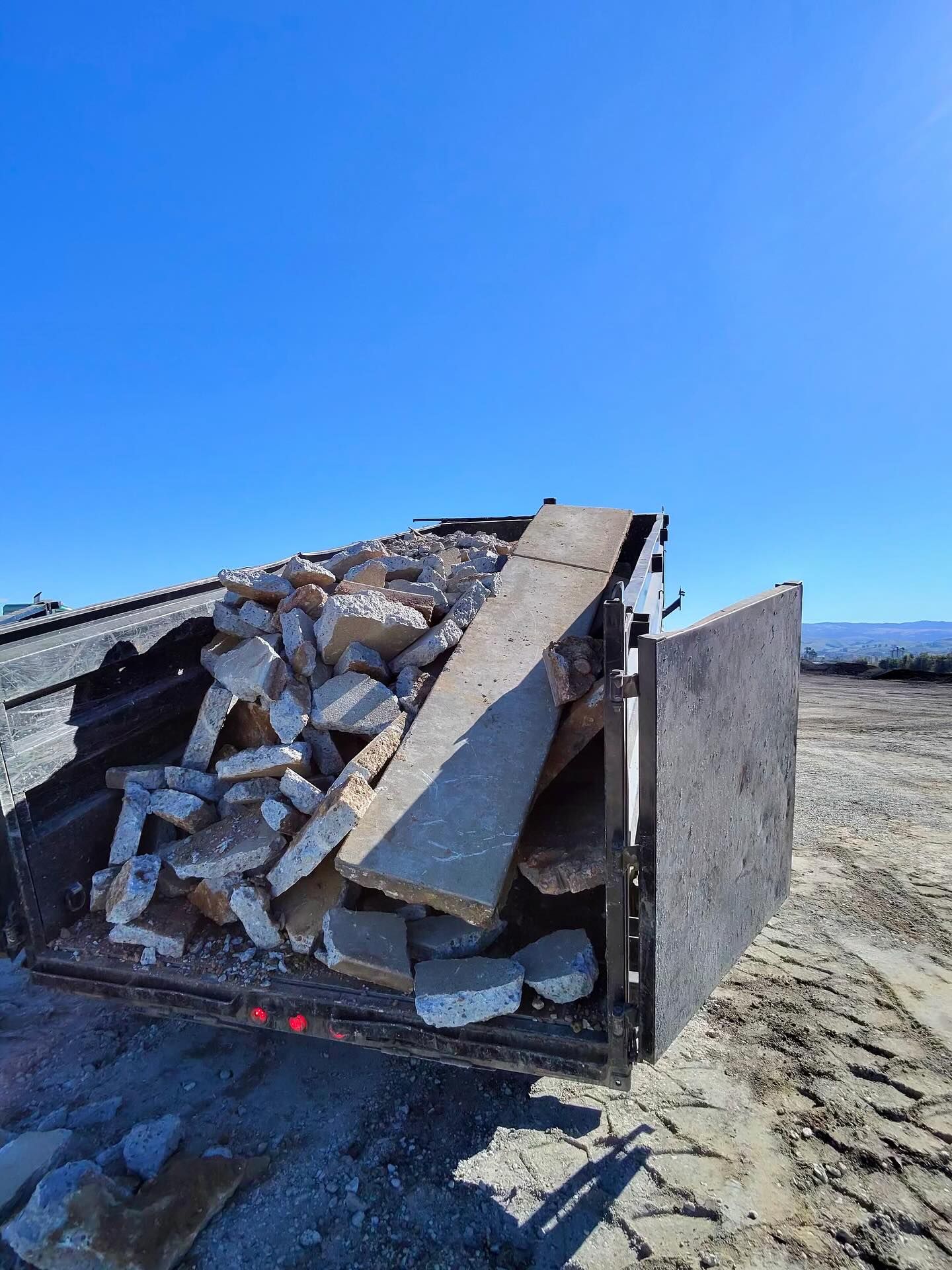 Dump trailer filled with broken concrete slabs and debris at a construction site, showcasing demolition waste removal, material hauling, and site cleanup under clear blue sky in an open landscape.