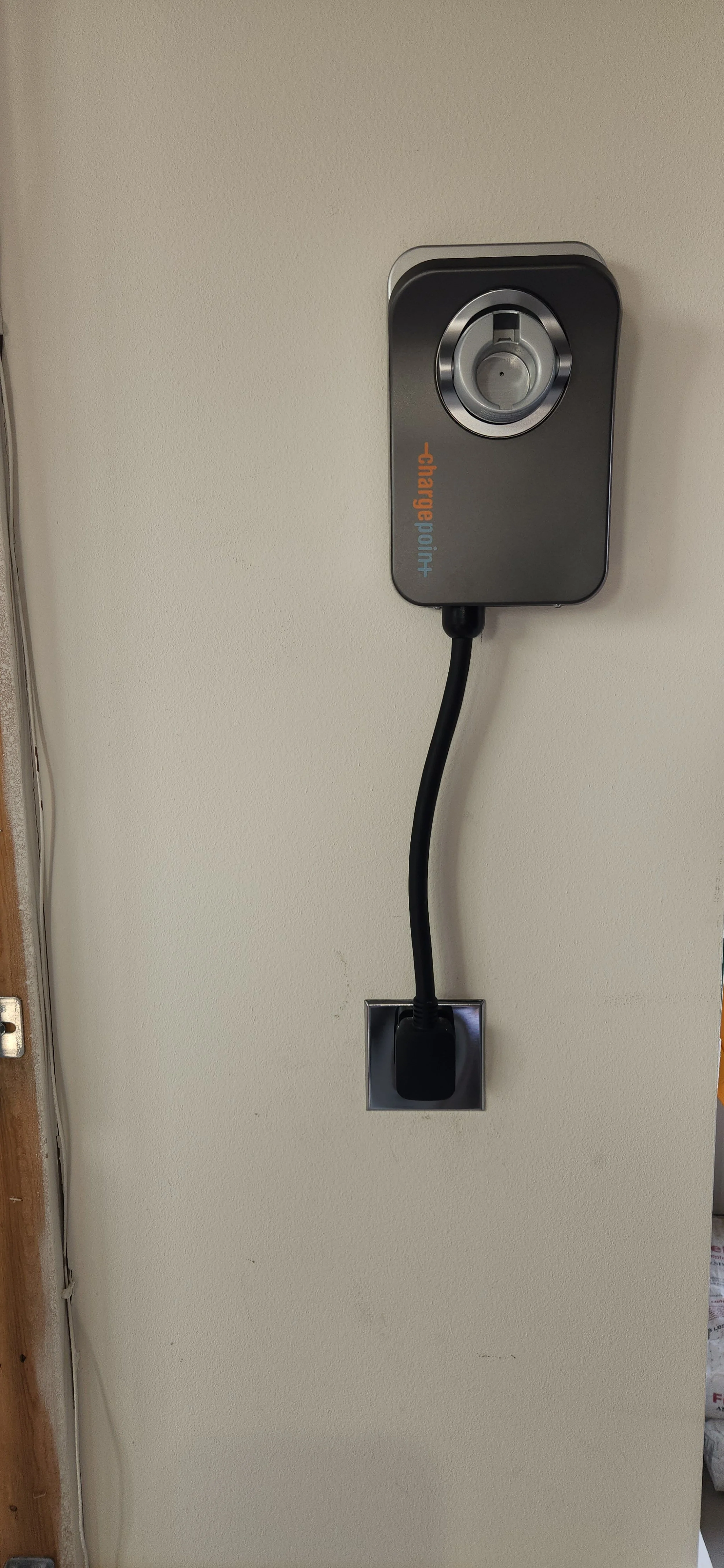 A ChargePoint electric vehicle charging station is mounted on a light-colored wall. A black charging cable extends from the station, connecting to a wall outlet below. The charging station is dark gray with a silver ring around the charging port and the ChargePoint logo is visible on the front.
