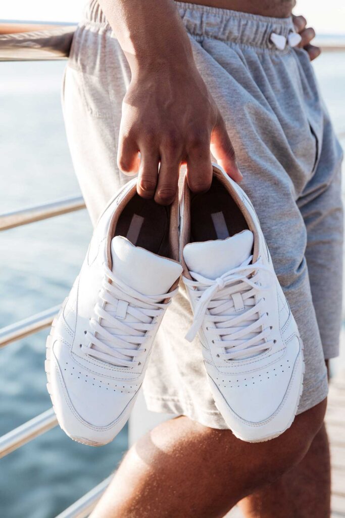 A person is holding a pair of white sneakers with grey shorts and a blurred ocean in the background.