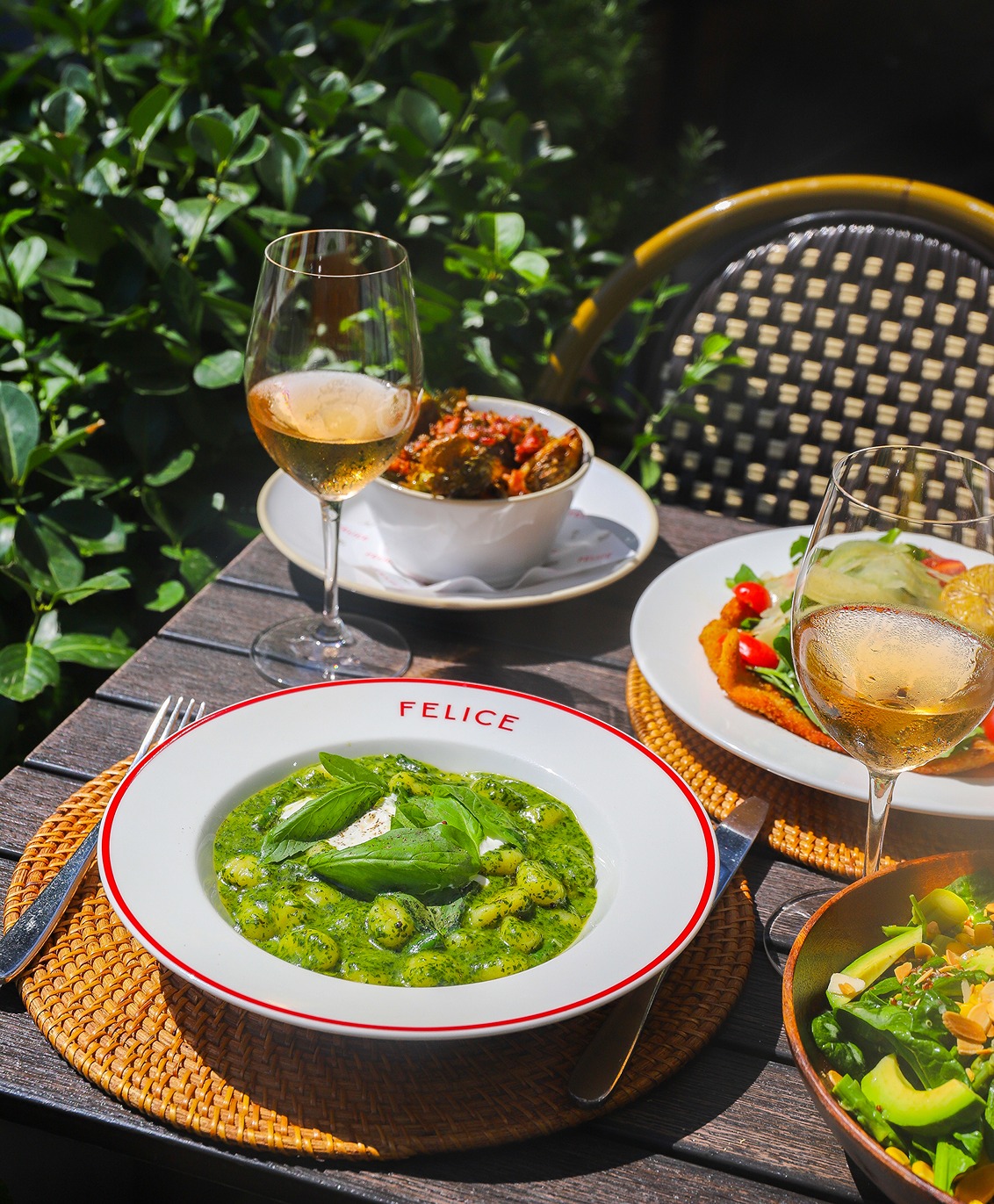 A plate of gnocchi in a bright green herb sauce topped with burrata and basil, served on a woven placemat with glasses of chilled white wine in a lush garden setting.