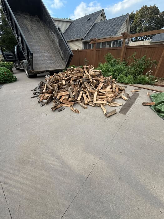 A work truck with its dump bed raised at a steep angle after delivering a large heap of split firewood directly onto a clean, light gray concrete driveway. A wooden privacy fence, green garden plants, and a residential house form the backdrop.
