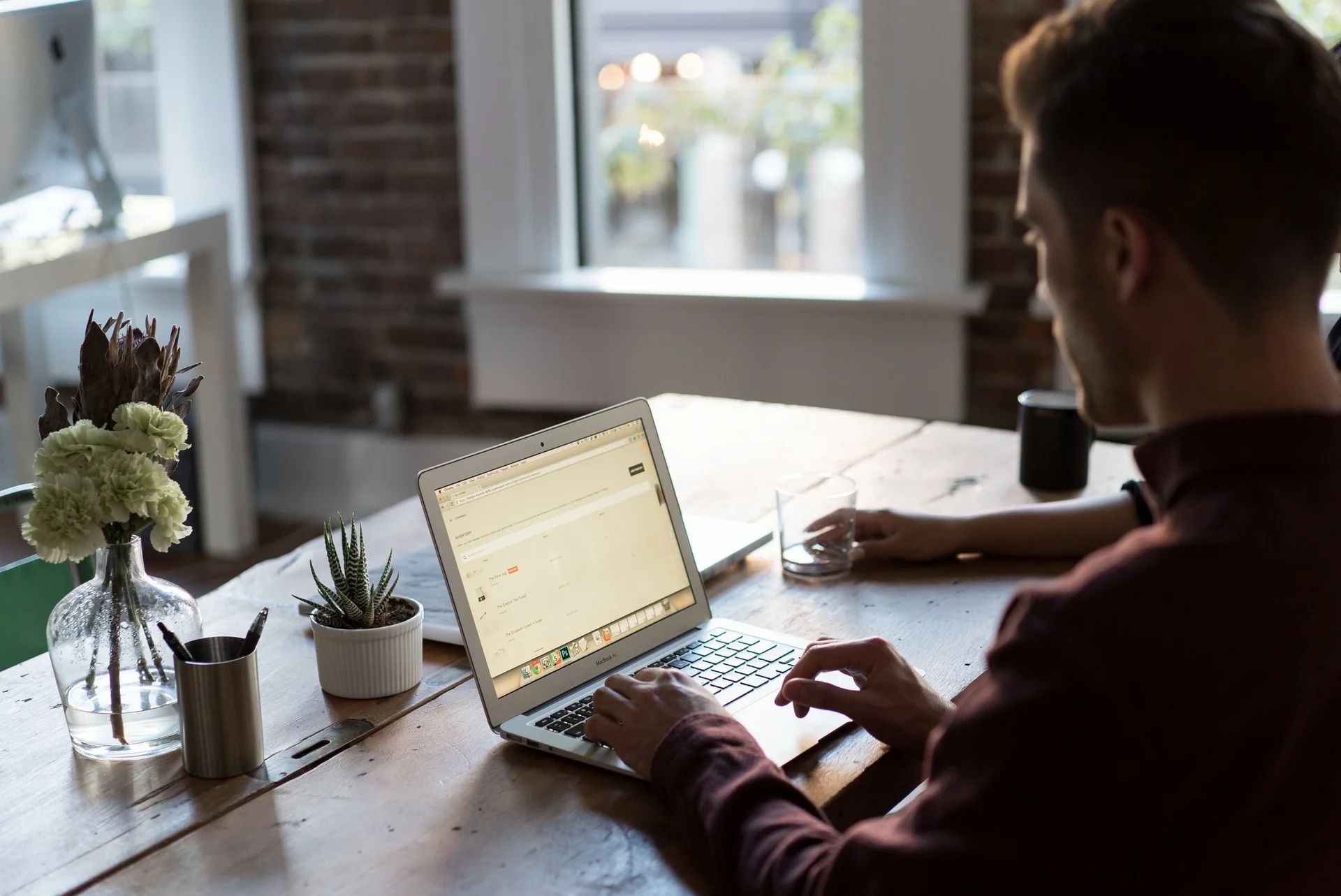 A man is typing on a laptop in a modern office or co-working space, with a vase of flowers and a small succulent on the table.