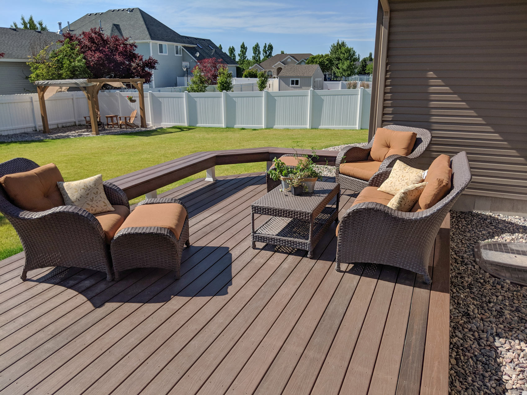Create the ultimate space for relaxing and entertaining with this custom composite deck by DeckPro LLC. This expansive deck features low-maintenance, dark brown composite decking boards and a unique built-in perimeter bench that doubles as safe railing. Furnished with comfortable wicker lounge seating and a coffee table, it&rsquo;s the perfect spot to enjoy your meticulously manicured lawn and peaceful suburban backyard.