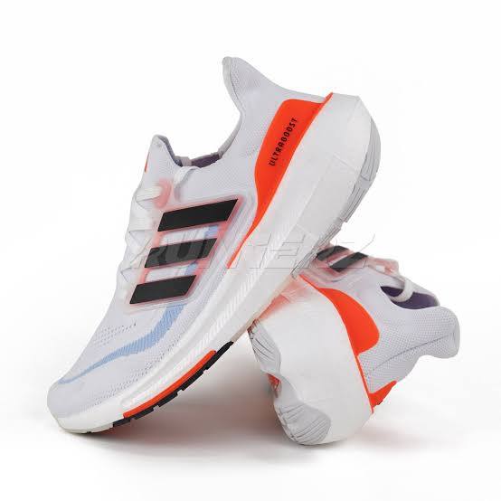 A pair of white and orange Adidas Ultraboost running shoes are shown against a white background. The shoes feature the signature Adidas three stripes in black on the side, and the word "ULTRABOOST" is printed vertically in orange on the heel. The sole of the shoes has some blue and orange accents.