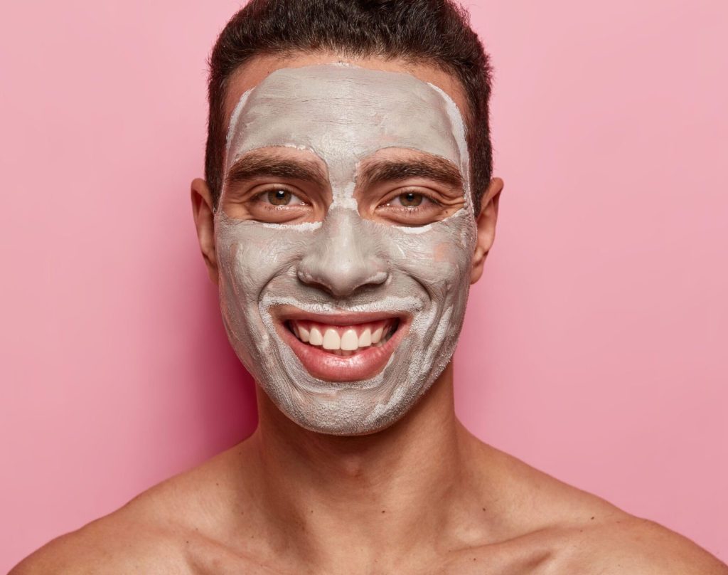 A man with short, dark hair is smiling widely while wearing a thick, grey clay mask on his face. The mask covers his forehead, cheeks, nose, and chin, leaving his eyes and mouth exposed. He is shirtless and his shoulders are visible. The background is a solid, bright pink color.
