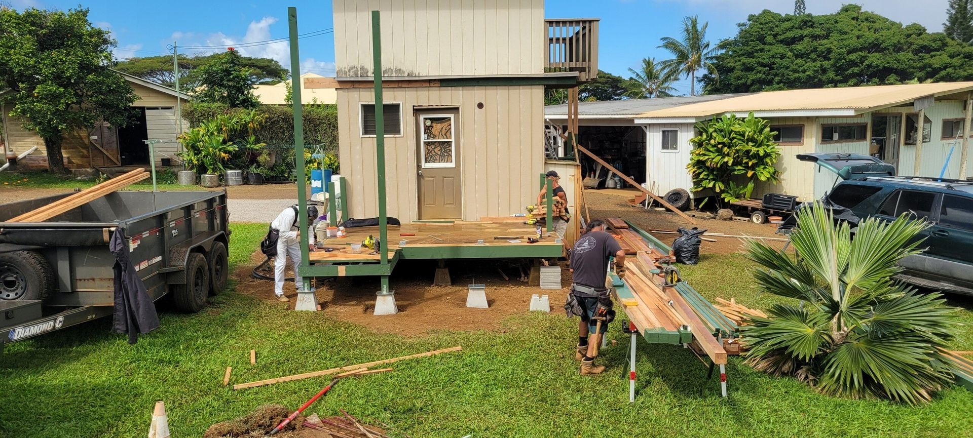 Construction workers are building a wooden deck onto a small, two-story building in a sunny, grassy yard. Lumber is being cut and installed, with a dump trailer visible nearby.