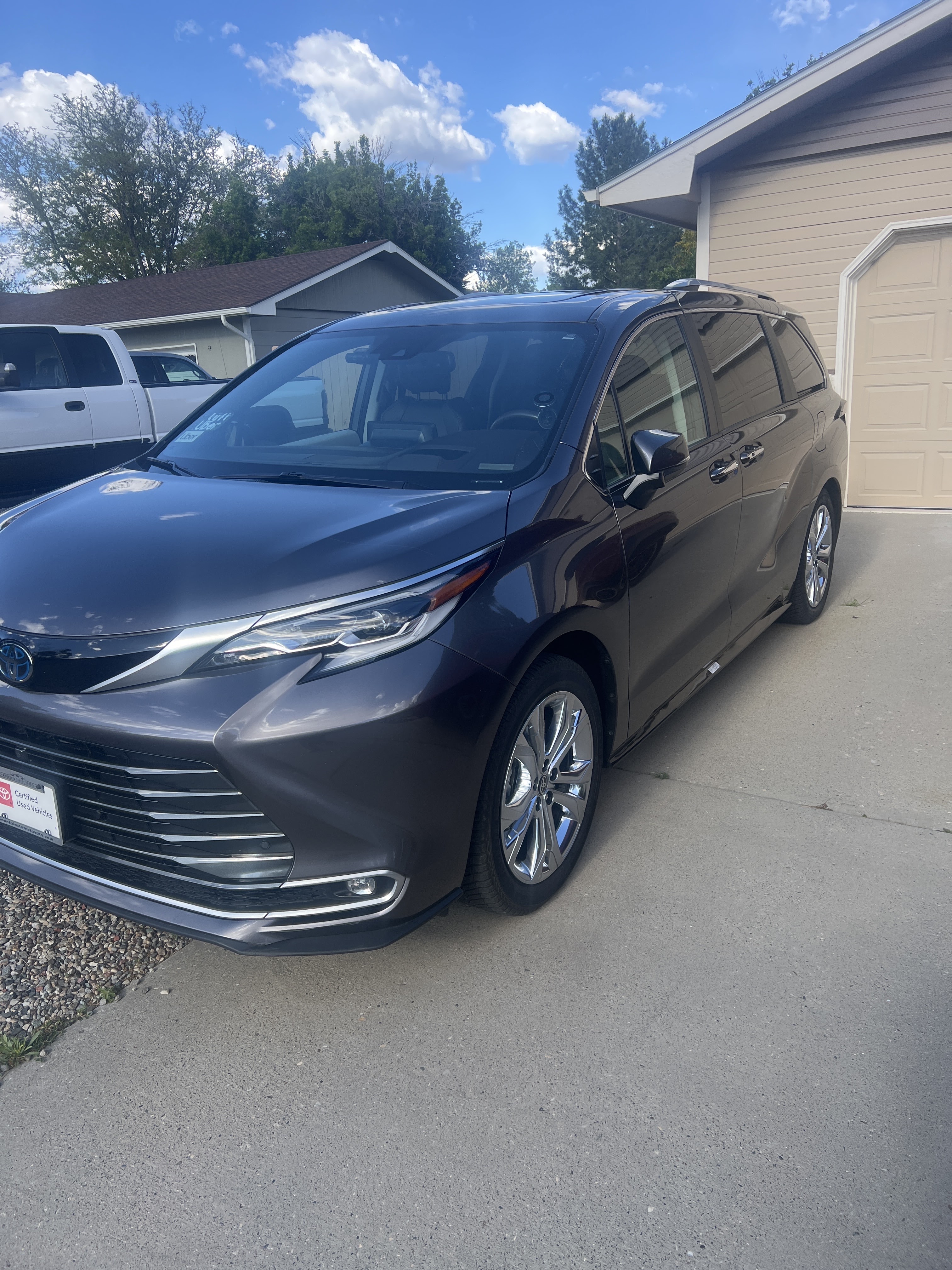 A dark gray Toyota Sienna minivan is parked in a driveway on a sunny day. The minivan has chrome wheels and a large grille. It is parked in front of a beige house with a garage door. There is a white pickup truck parked to the left of the minivan. The sky is blue with white clouds.