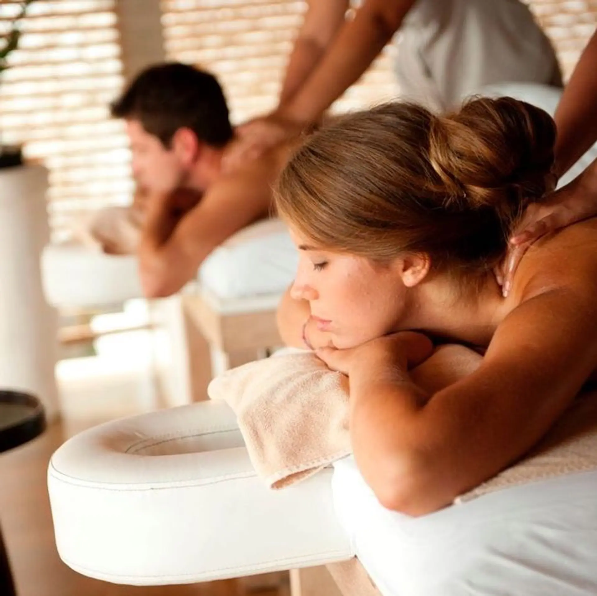 A close-up shot of a woman receiving a massage, with a man in the background also receiving a massage. Both individuals are lying on massage tables, and their backs are being attended to by massage therapists. The setting appears to be a spa or wellness center, with a tranquil and relaxing ambiance. The lighting is soft and warm, enhancing the serene atmosphere.