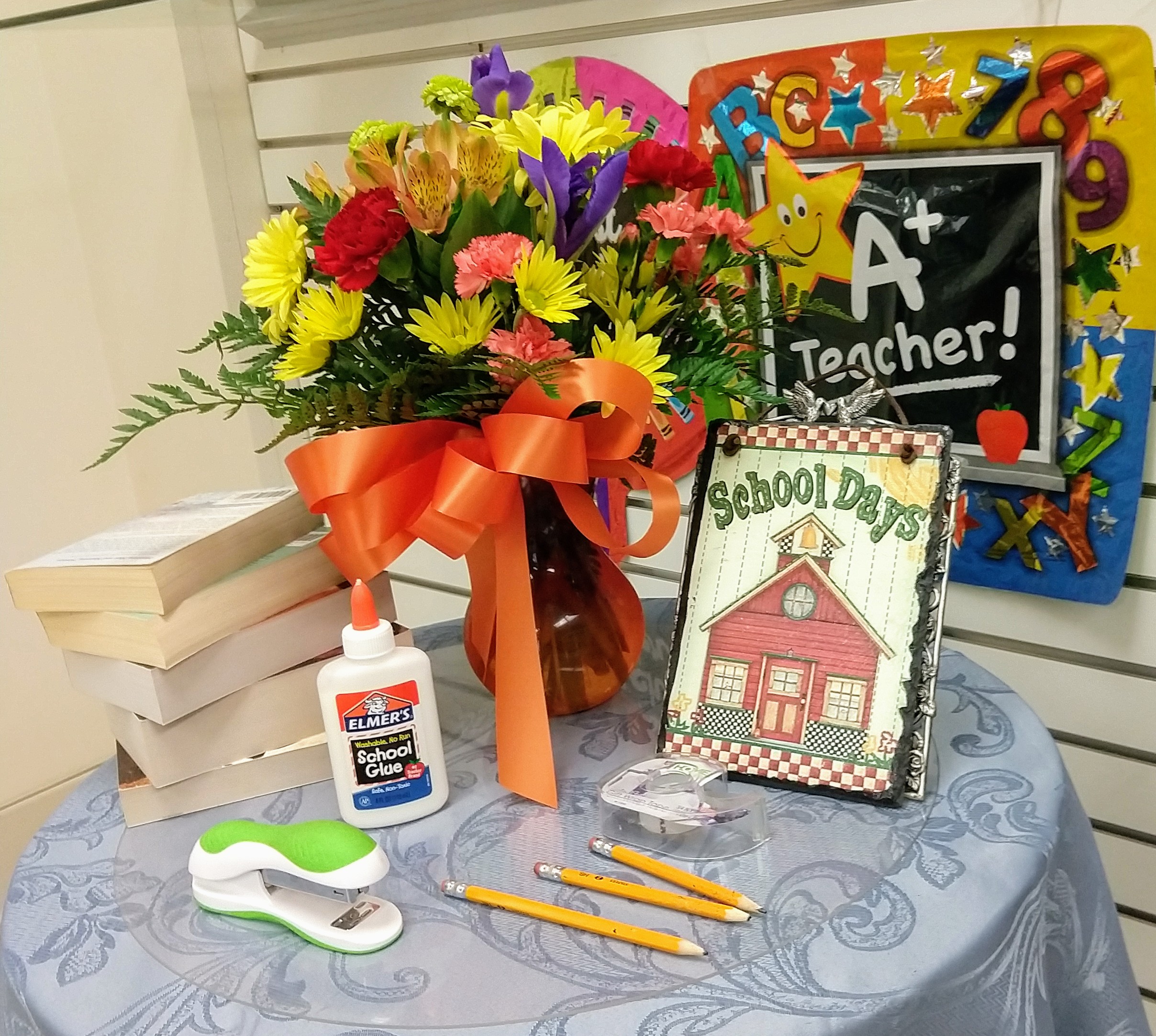 Fort Bragg Flower Shop back to school display table with a vase of fresh flowers including yellow daisies, blue iris, red carnations, orange spray roses, and a giant orange bow.