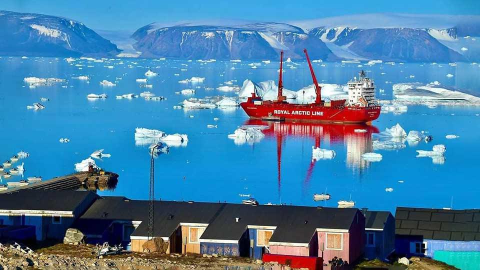 A small Greenland fishing and shipping village in the foreground, blue water with ice flows and mountains in the middle and background. A red shipping container sails in the middle of the picture, with 2 tall cranes on deck.