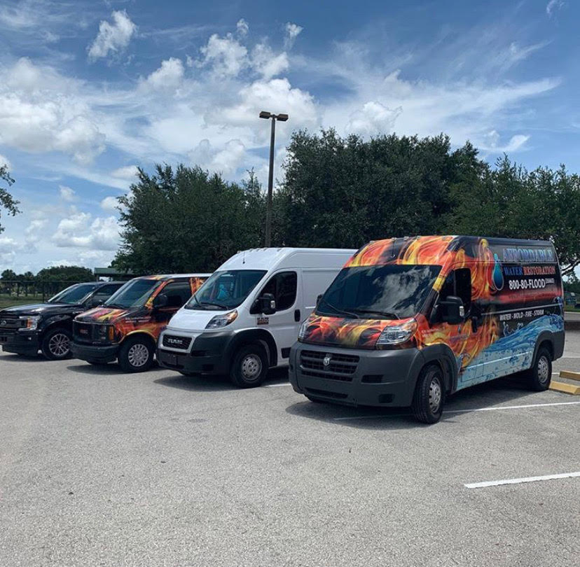 A line of service vans are parked in a lot under a partly cloudy sky. The vans are decorated with graphics related to fire and water. The front van has a logo and contact information for "Affordable Water Restoration" with a phone number and services listed. The vans are parked in a paved lot with some trees and a lamppost visible in the background.