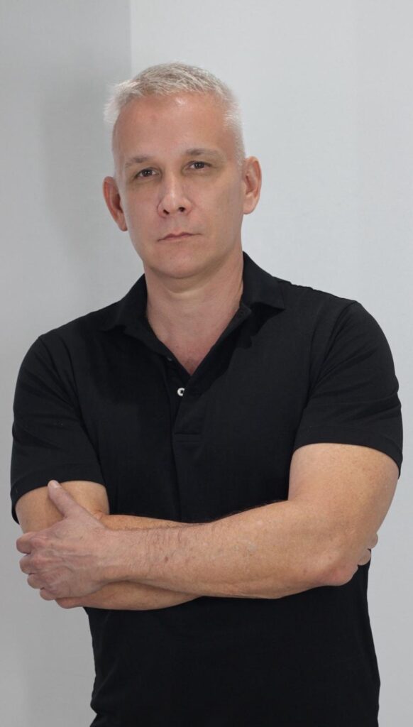 A medium shot of a fair-skinned man with short, gray hair and a black collared shirt standing in front of a plain white background. His arms are crossed, and he is looking directly at the camera with a neutral expression. The lighting is bright and even, highlighting the man's features and the texture of his shirt.