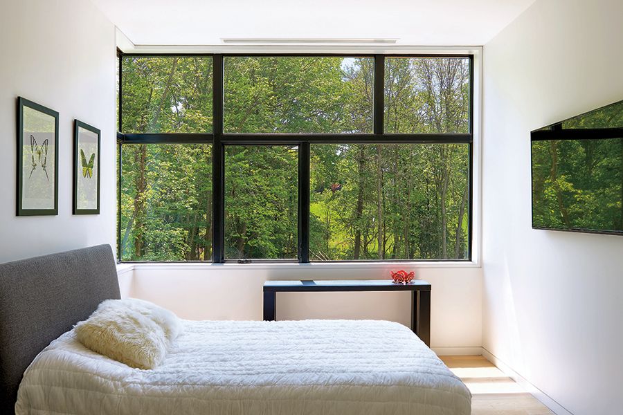 Large black-framed picture windows fill a bright bedroom with natural light, offering a wide view of green trees outside.