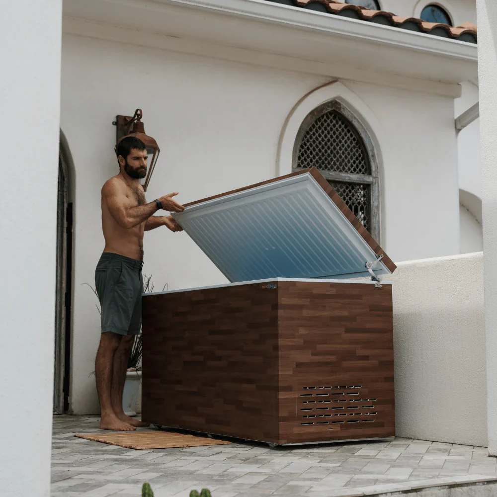 A shirtless man with a beard is standing next to a large, wooden chest freezer, holding the lid open. The freezer is located outdoors on a tiled patio, with a white building and an arched window in the background. The lid of the freezer appears to be filled with ice or water.