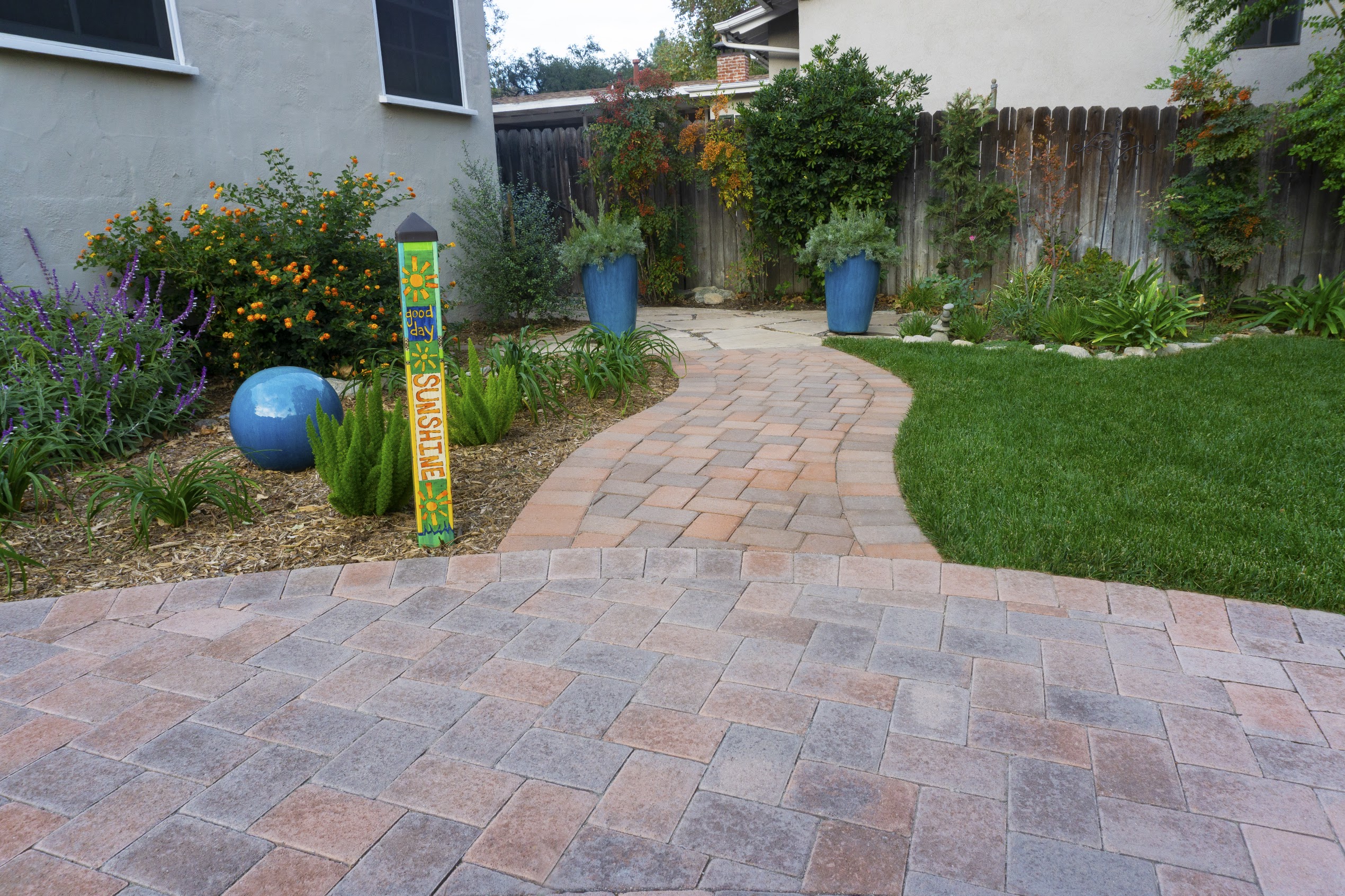 Curved brick paver walkway winds through a colorful backyard garden with flowers, blue pots, green lawn, and a bright welcome sign.
