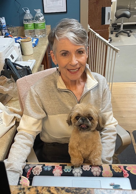 A woman with short gray hair is sitting at a desk with a small, fluffy tan dog sitting on her lap. She is smiling at the camera. In the background, there are bottles of water, a printer, a handbag, and a baby gate. The woman is wearing a gray and white zip-up sweatshirt. The dog is looking at the camera with its tongue sticking out slightly. The desk has a colorful mousepad with paw prints on it.