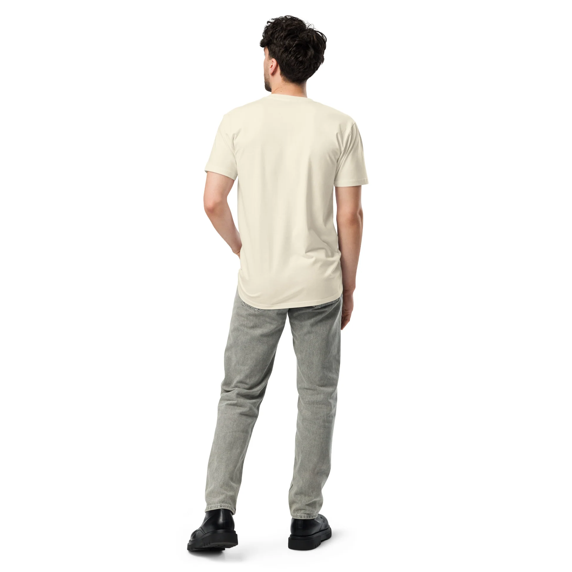 A man stands with his back to the camera, wearing a light yellow t-shirt and grey jeans. He has his left hand in his pocket. The background is plain white.