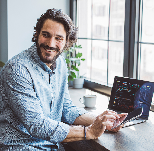 A man smiling while reviewing financial data on both his laptop and tablet.
