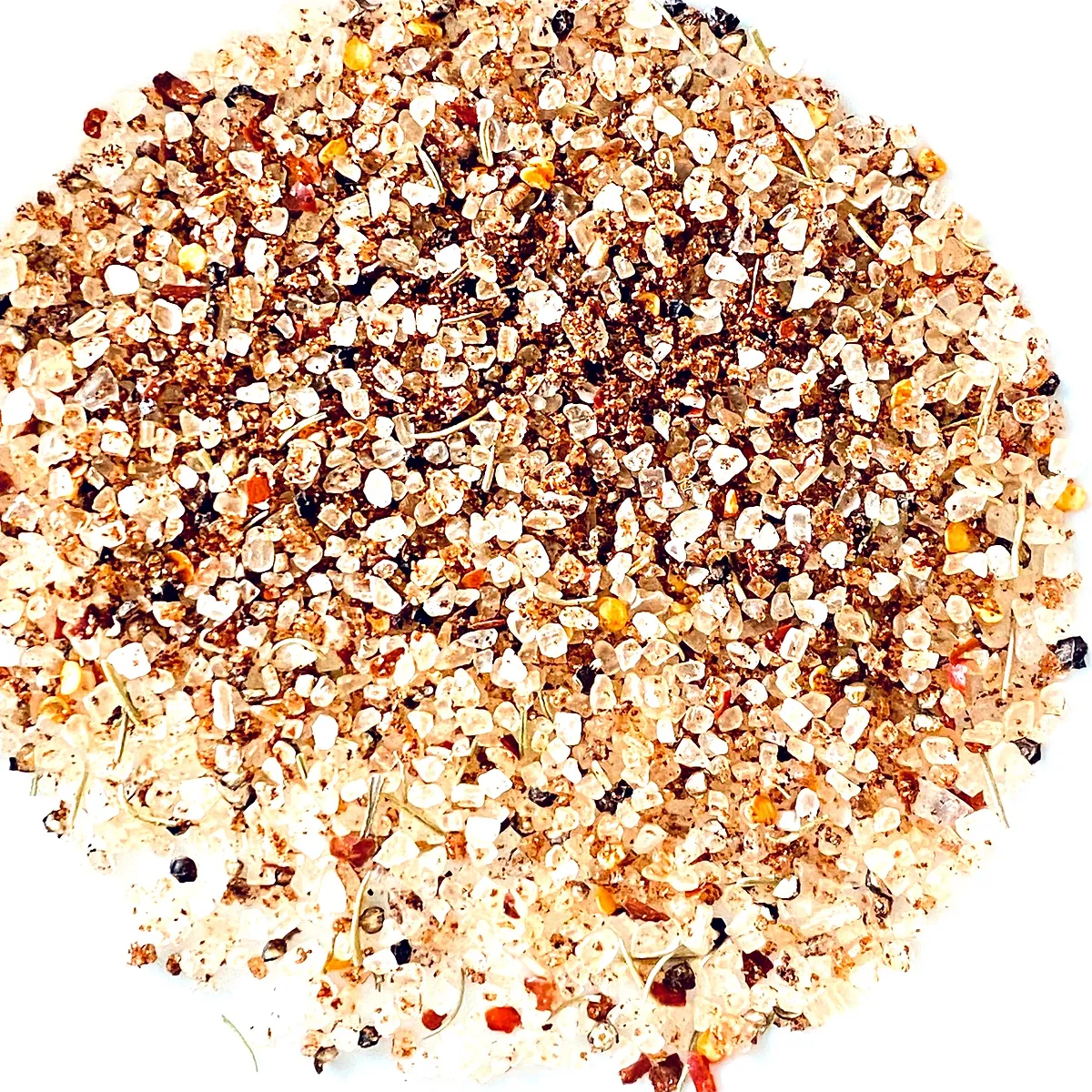 A top-down, close-up shot of a pile of mixed spices and salt. The texture is granular with visible pieces of salt crystals, herbs, and possibly small dried peppers or seeds. The pile is circular and against a white background.