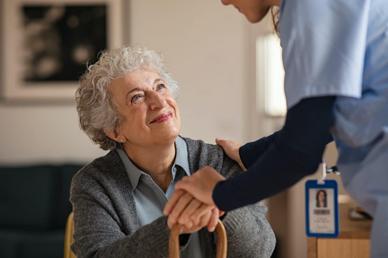 A close-up of an elderly woman with curly gray hair looking up with a smile, as a caregiver in a light blue uniform places a hand on her shoulder and another hand on her clasped hands, which rest on a cane. The caregiver also has a blue ID badge clipped to their uniform. The background appears to be a room with a picture on the wall and some furniture.