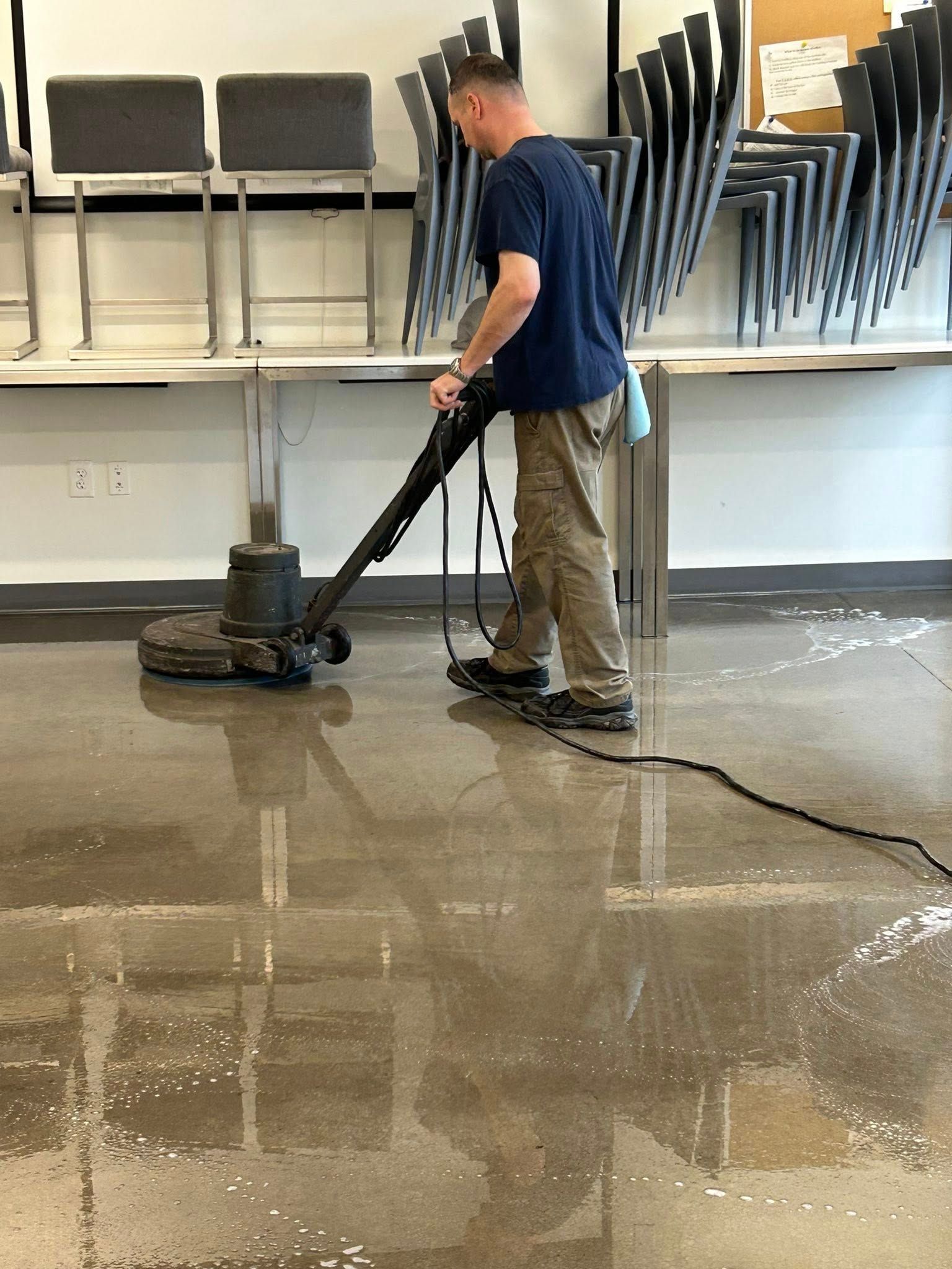 Janitorial worker using floor buffer machine to clean and polish commercial floor, removing dirt and restoring shine in indoor space with stacked chairs, demonstrating professional cleaning and maintenance services.