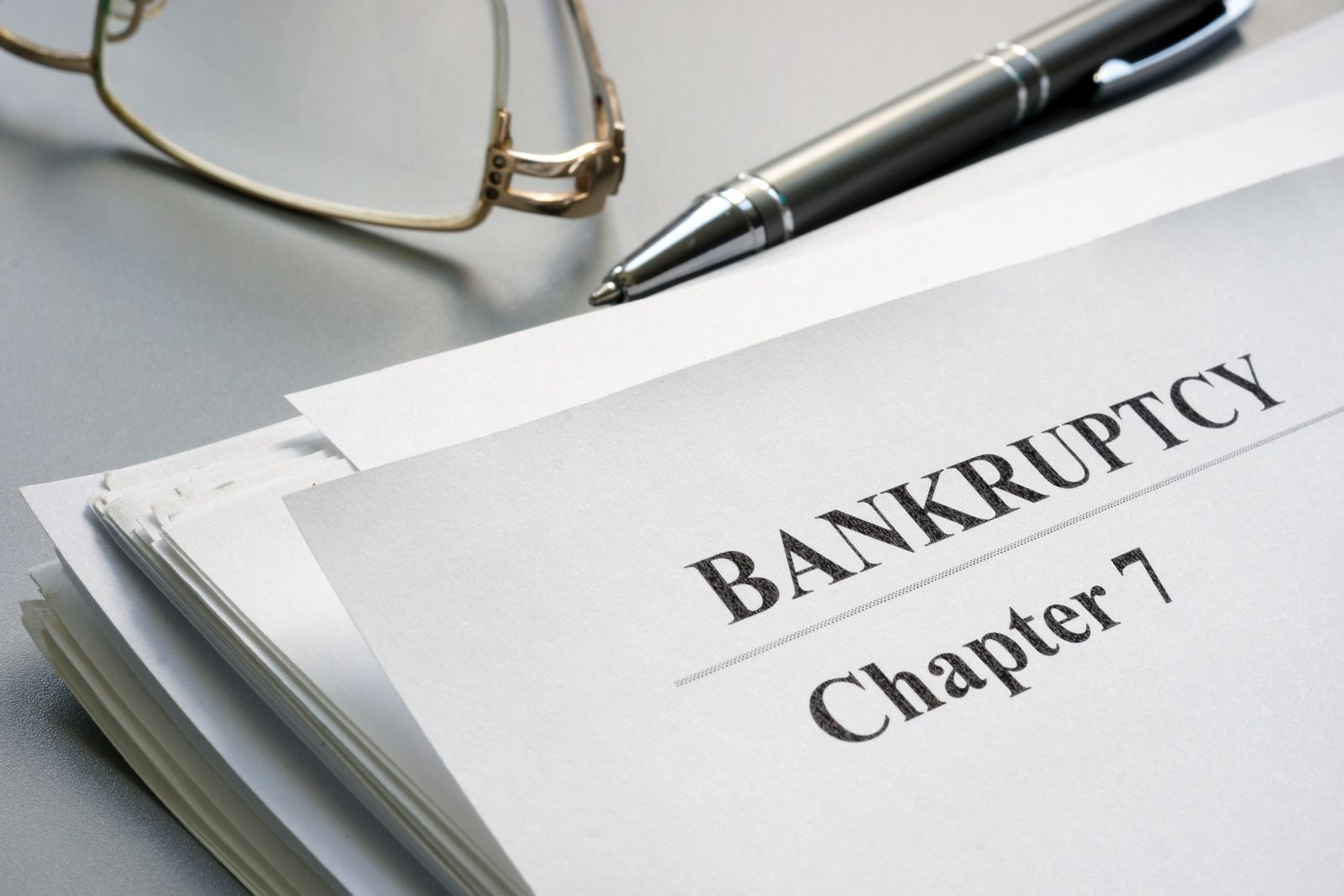 A stack of papers headed "Bankruptcy Chapter 7" sits on a gray desk, with a pair of gold-framed eyeglasses and a dark metallic ballpoint pen resting behind the documents.