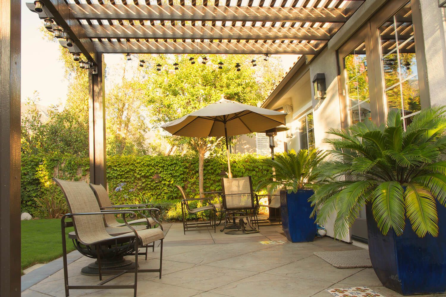 Sunlit backyard patio with pergola, lounge chairs, umbrella table, lush palms, and greenery, creating a warm, peaceful outdoor retreat.