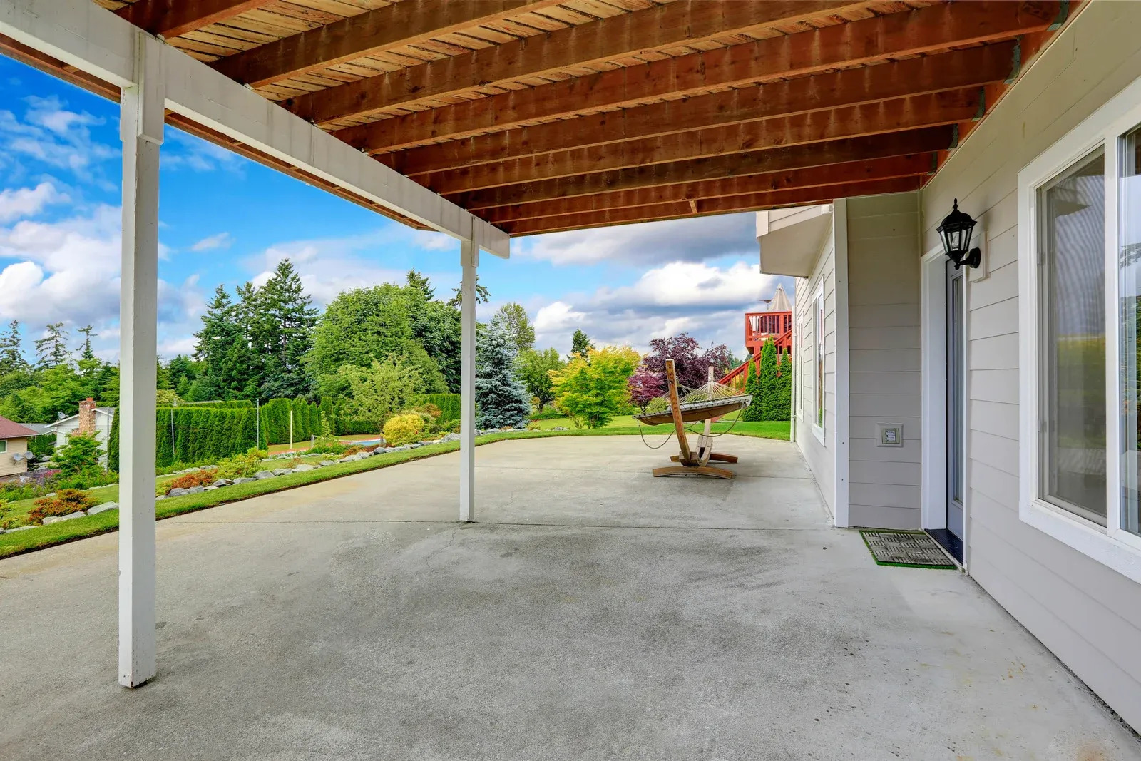 A spacious concrete patio with a wooden pergola extends from a house, offering an open-air area with views of a lush backyard under a partly cloudy sky.