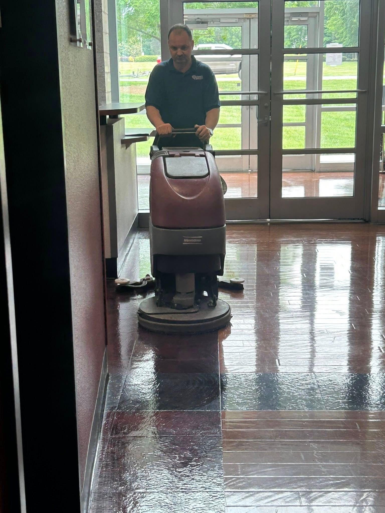 Professional cleaner operating a floor scrubber machine inside commercial building entrance, polishing hard floors to a glossy finish, demonstrating routine janitorial maintenance and cleanliness in a high-traffic indoor environment.