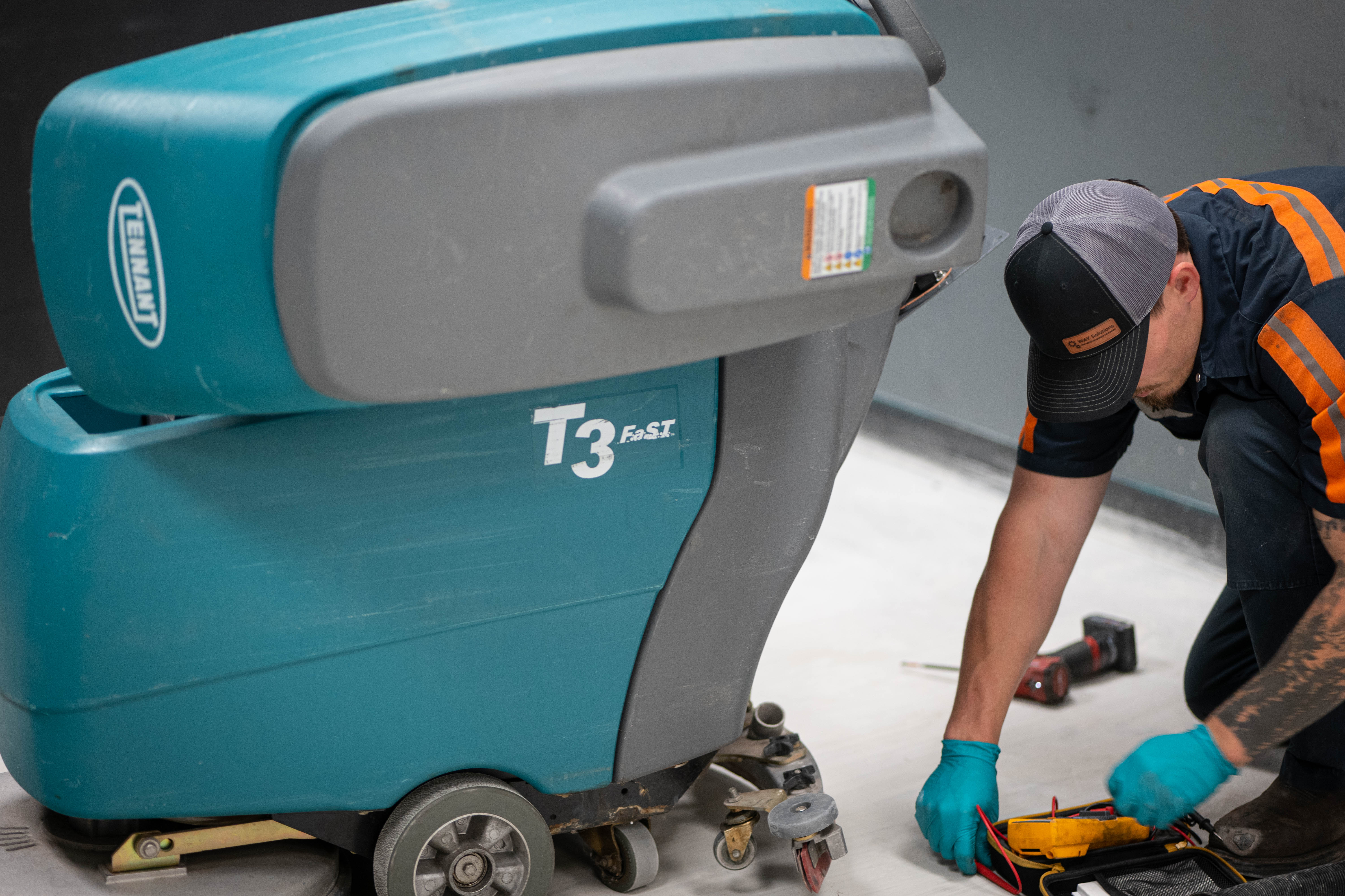A certified WaySolutions technician prepares a digital multimeter to perform data-driven electrical diagnostics on a teal Tennant T3 FAST walk-behind floor scrubber in Layton, Utah. Our precision testing ensures every circuit and relay in your commercial cleaning equipment operates flawlessly.