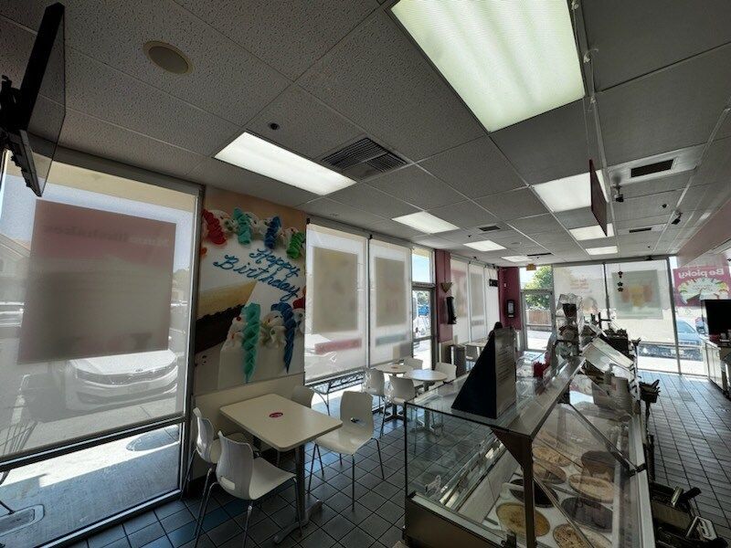 A bright, clean ice cream parlor interior showcasing white roller shades that diffuse natural light. The space includes modern seating, a tiled floor, and a colorful birthday-themed wall graphic.