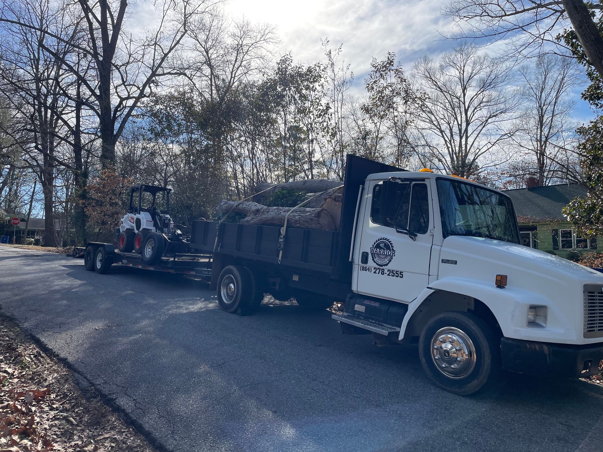 Tree service truck and trailer parked on a residential street carrying large cut logs and a skid steer, indicating active tree removal or hauling operations in a neighborhood setting.