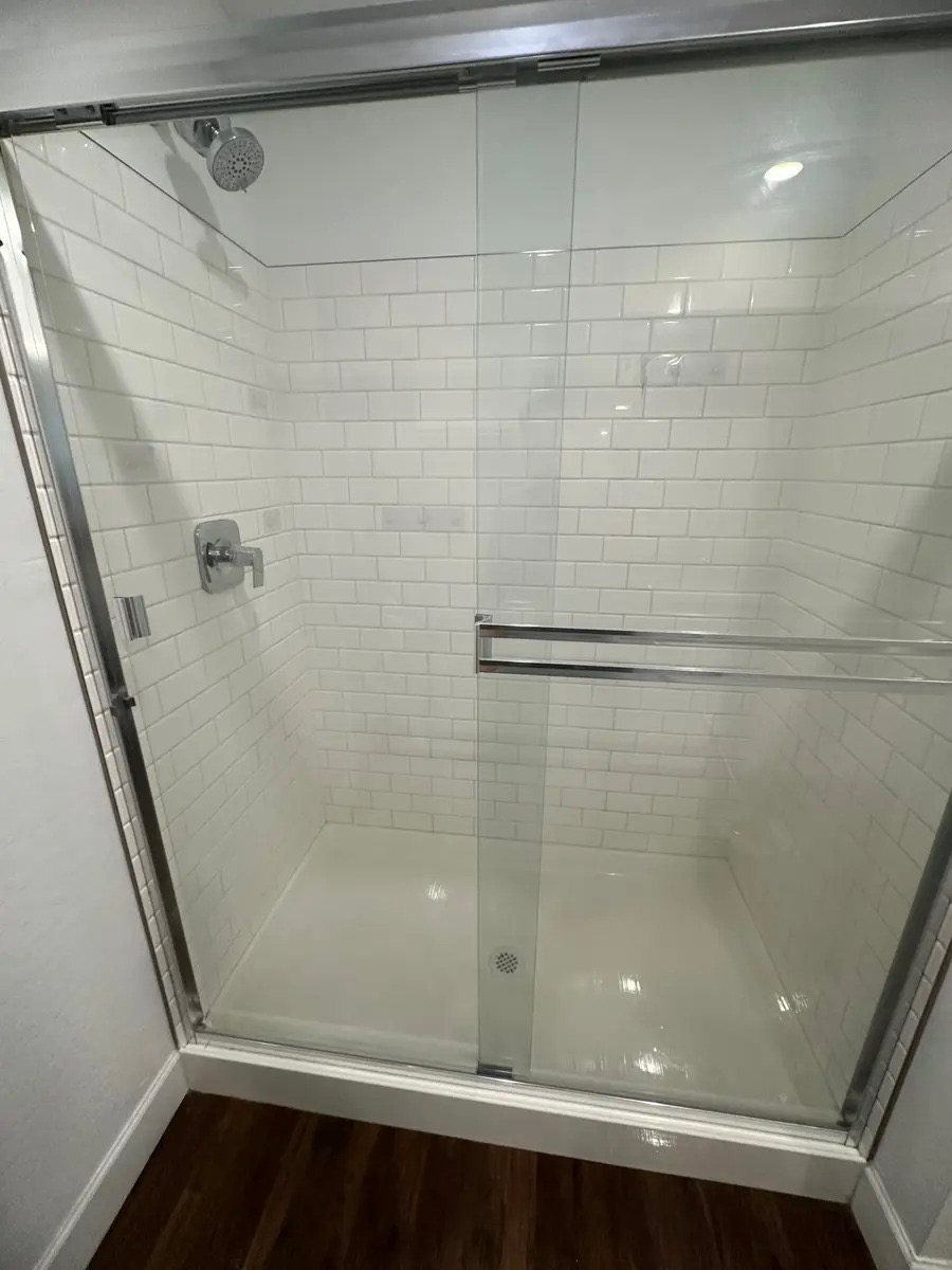 A modern bathroom shower with white subway tile walls, a glass sliding door with a chrome handle, a chrome showerhead and faucet, and a white shower base with a drain. The floor outside the shower is dark wood. The shower appears to be very clean and well-maintained.
